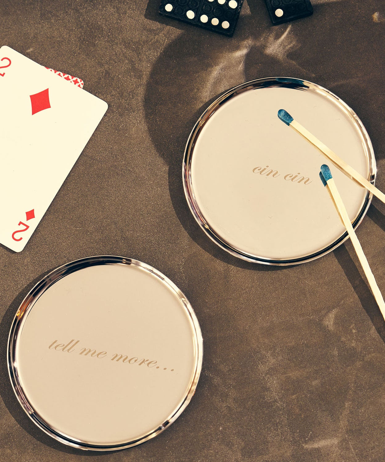 Two round silver-framed coaster with re arranged on a textured brown tabletop. One mirror has the phrase 'tell mel me more', the other has 'cin cin'