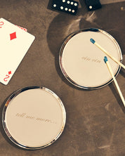 Two round silver-framed coaster with re arranged on a textured brown tabletop. One mirror has the phrase 'tell mel me more', the other has 'cin cin'